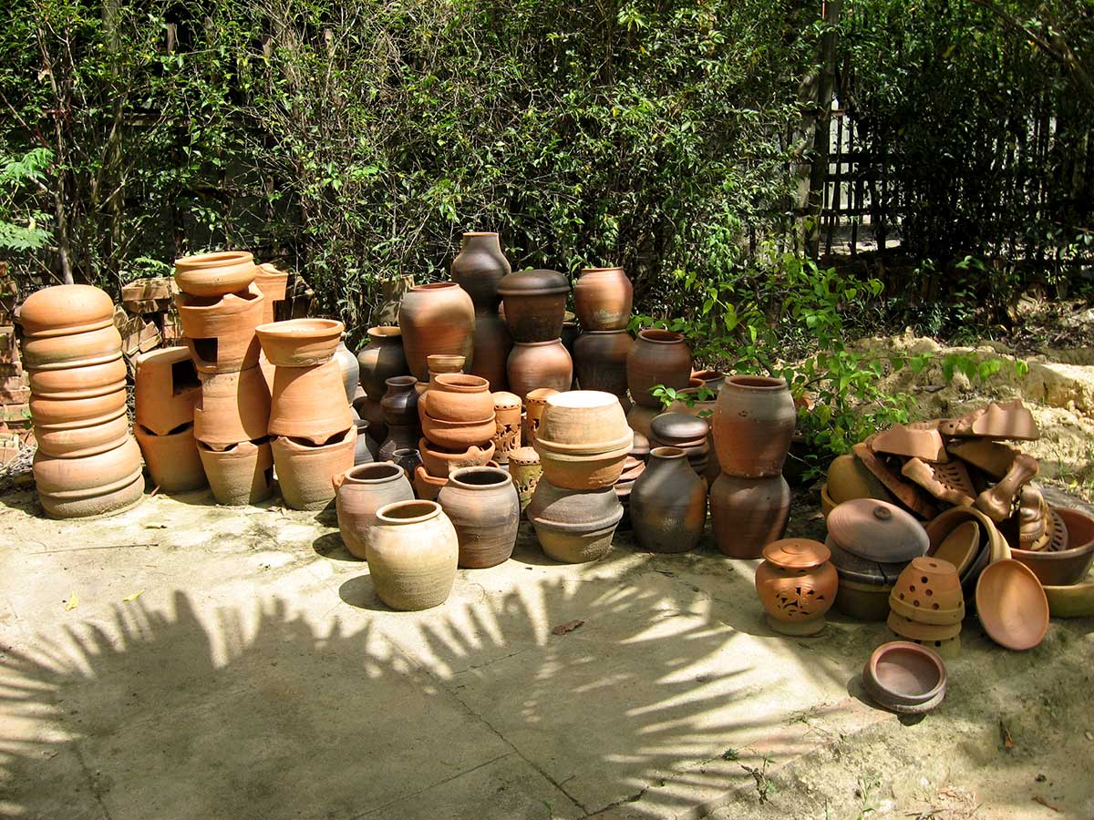 Dozens of unglazed red and brown earthenware jars, pots and vases of different shapes and sizes clustered together in a shaded garden, a few broken pieces and lids scattered among them.
