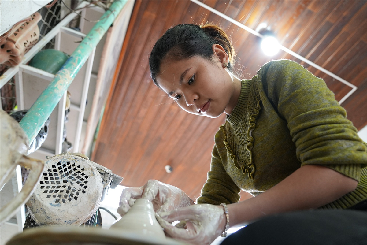 A young woman potter in Thanh Hà bends over a spinning wheel, shaping the neck of a clay vessel with both hands, wooden roof beams and shelved pottery visible in the workshop behind her.