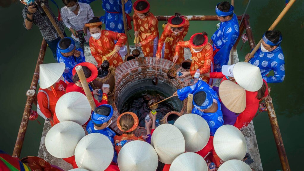 A bird's-eye view of an old brick-lined well: a crowd in red and blue áo dài, wearing conical hats and turbans, gathered around the mouth of the well drawing water with dippers and earthenware jars.