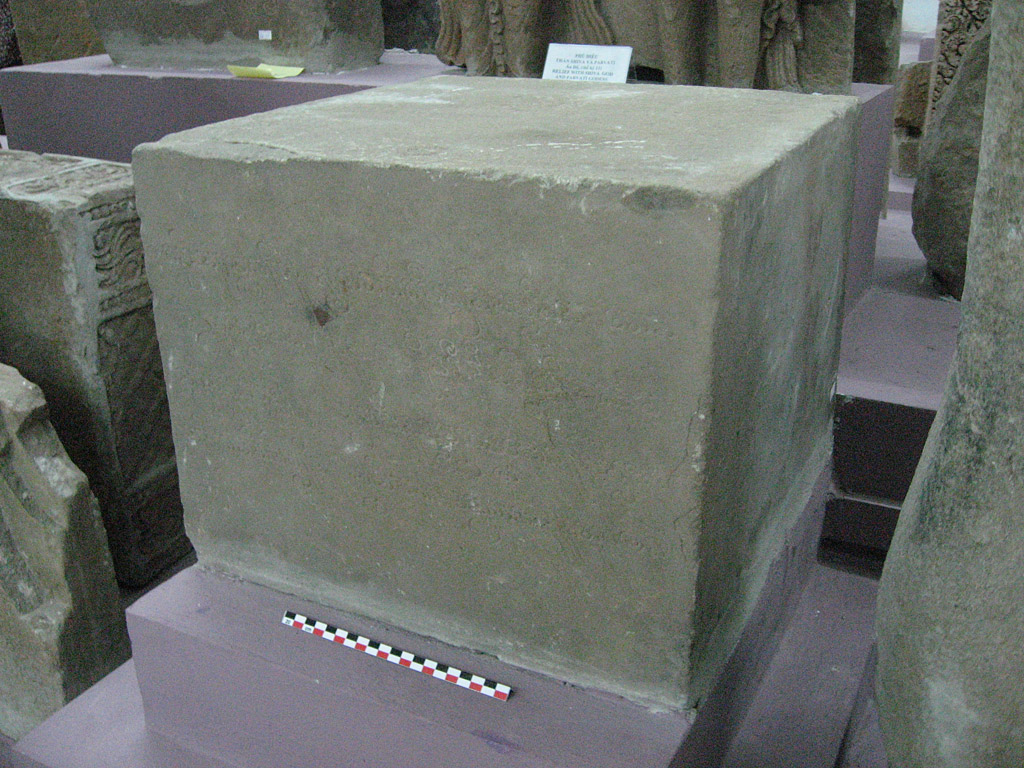 A square stone pedestal carved with faint Sanskrit script, inscription C.137 from Trà Kiệu, photographed in a museum storeroom in Hà Nội among other Champa stone fragments.