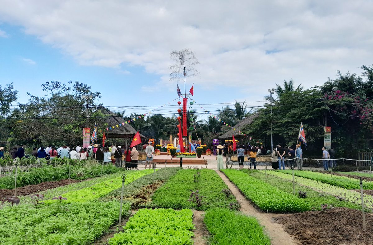 A tall New Year pole stands in the village yard hung with red streamers and lanterns, a red flag with a yellow star beside it. Onlookers stand behind and vegetable beds in several shades of green lie in front.