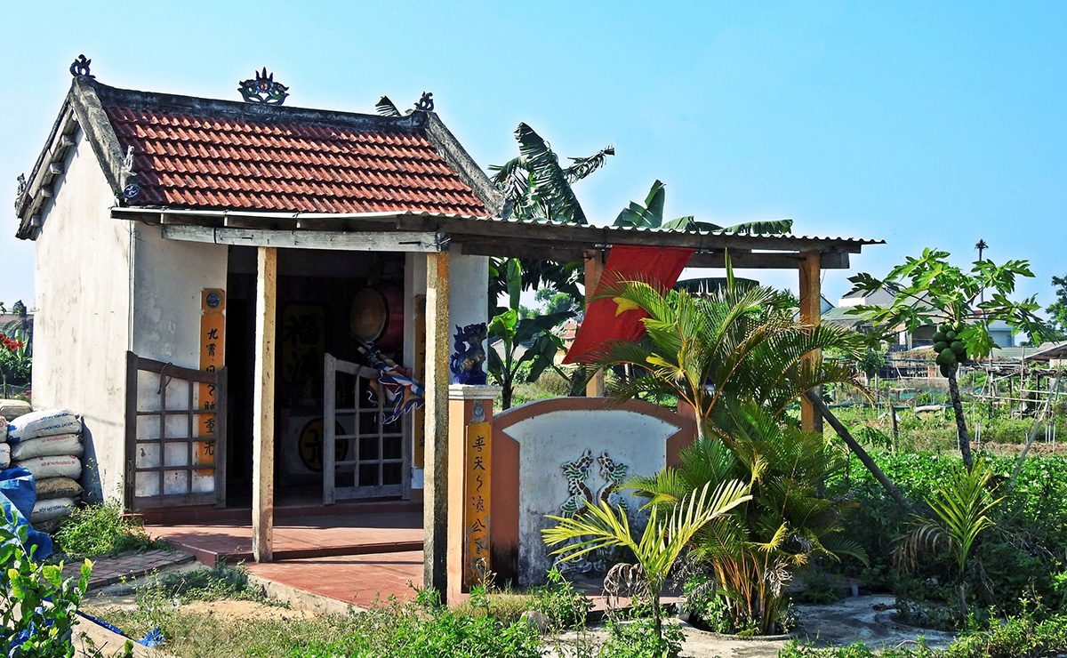 A small whitewashed shrine with a red-tiled roof and an open porch with two wooden pillars. In front, a relief screen wall, ornamental plants and bananas, and a red flag hanging from the porch. Sacks are stacked at left and a garden lies behind.