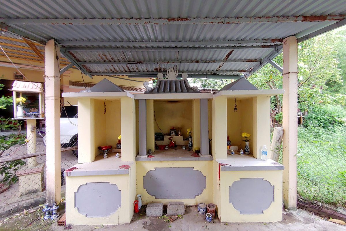 Three shrine niches built side by side, painted pale yellow, under one tin roof. The middle niche has a small roof and the two outer ones are lower. Each holds a vase of flowers and small offerings.