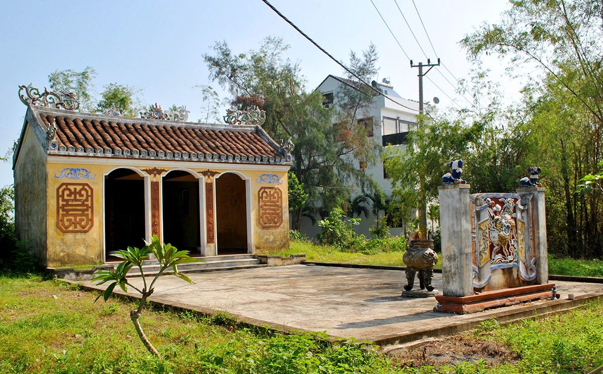 A yellow-walled shrine with a yin-yang tile roof and a three-arched front. Round relief patterns flank the doors. A relief screen wall and a large incense burner stand in the yard, with dense trees behind.